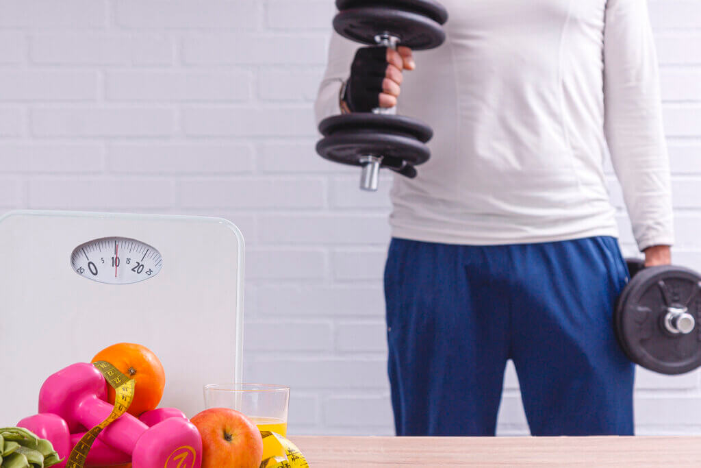 Hombre haciendo ejercicios con mancuernas y en primer plano báscula de peso, frutas, verduras y cinta métrica. concepto de dieta y deporte.