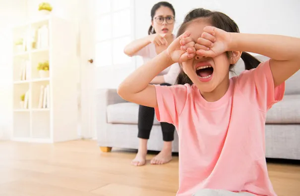 Niña llorando con su madre detrás regañando.