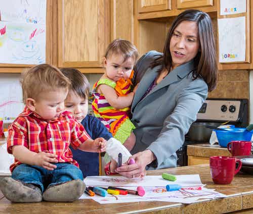 Mujer en la cocina con una niña en brazos y los otros dos hijos pintando en la cocina