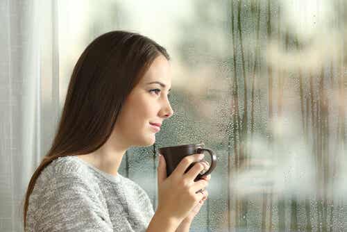 Femme qui regarde par la fenêtre avec une tasse dans les mains.