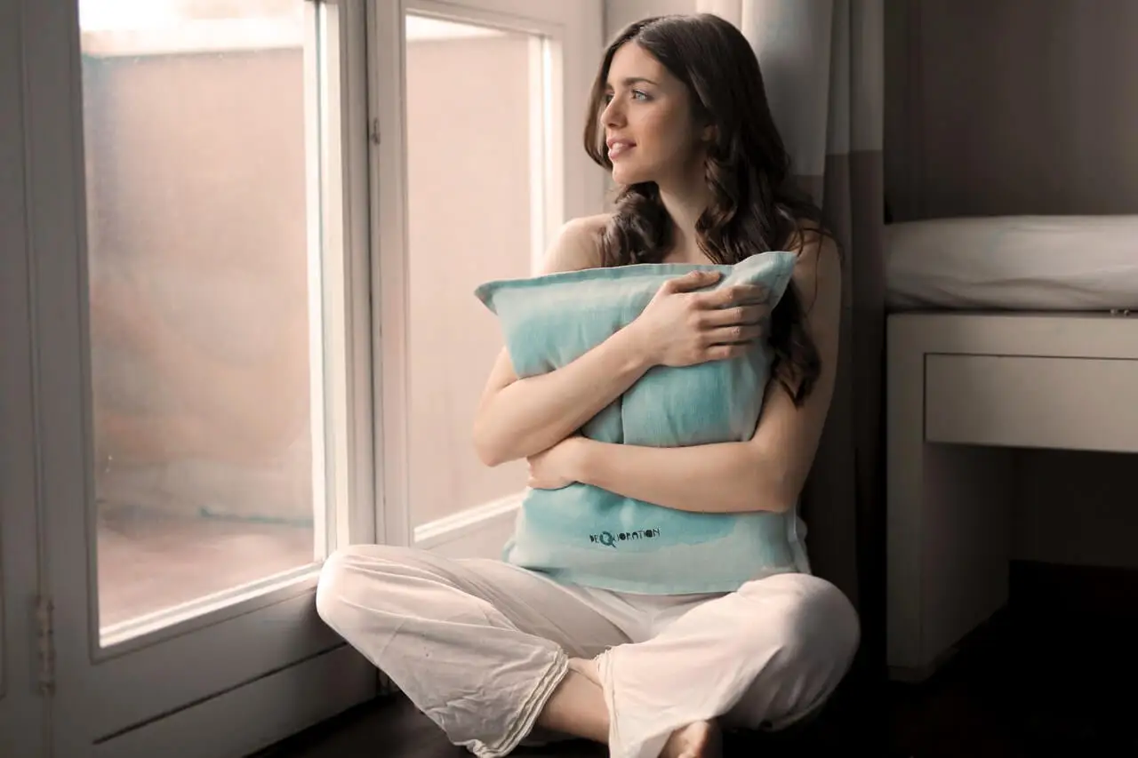 Mujer abrazando una almohada mirando por la ventana.