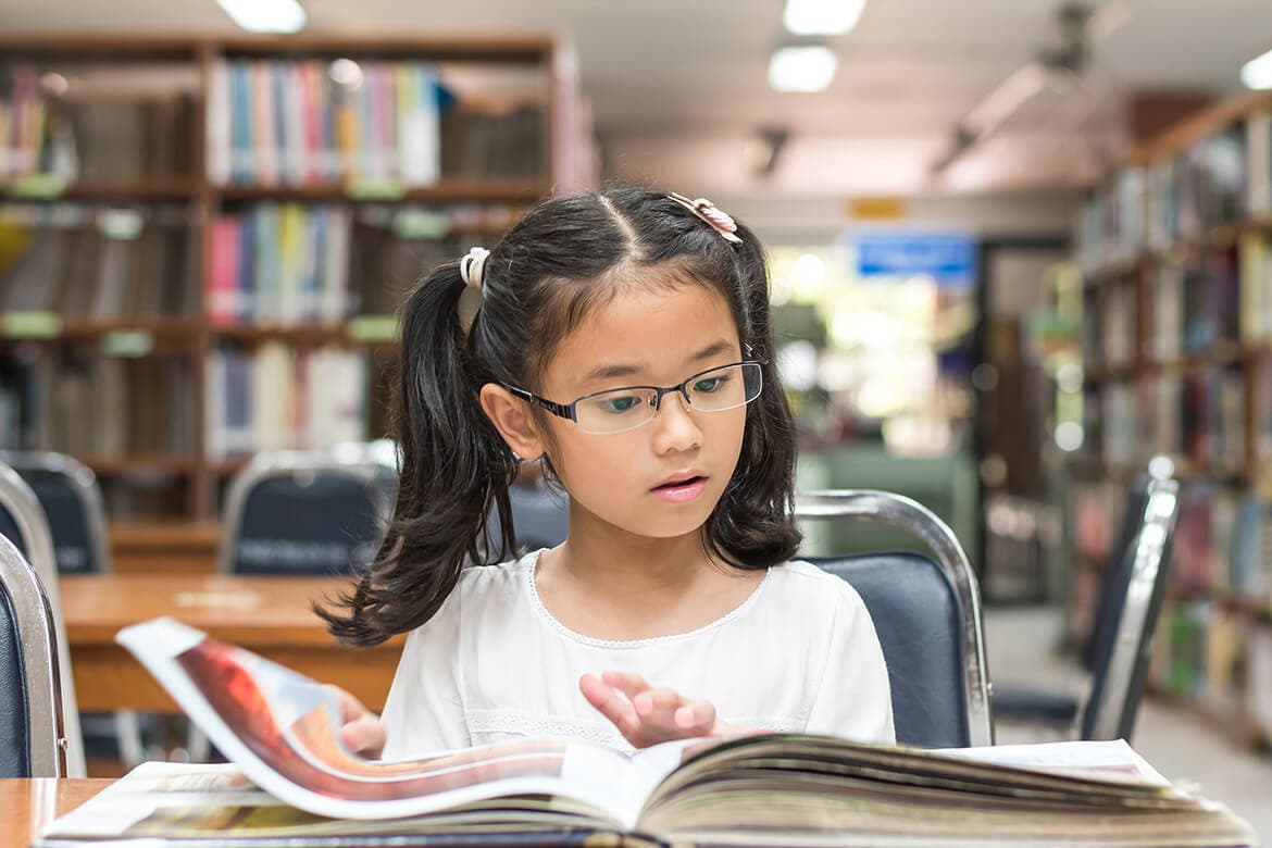 Niña pequeña lee un libro con fotografías en una biblioteca.