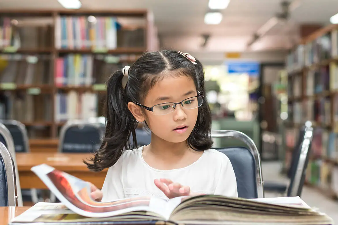 Niña pequeña lee un libro con fotografías en una biblioteca.