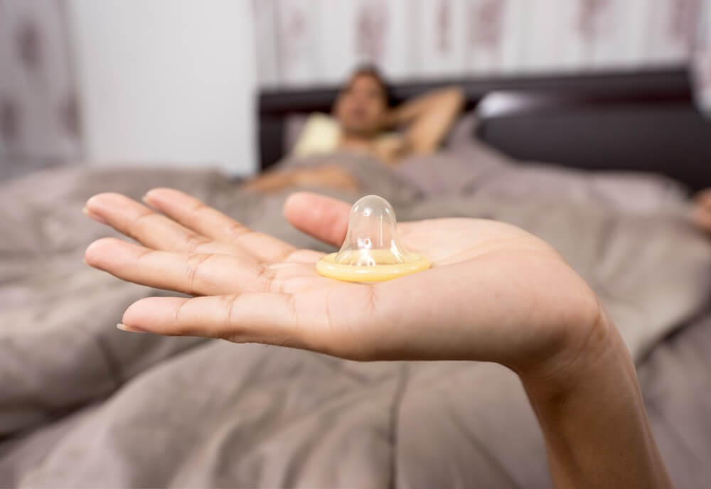 Mano sujetando un preservativo con el hombre en la cama