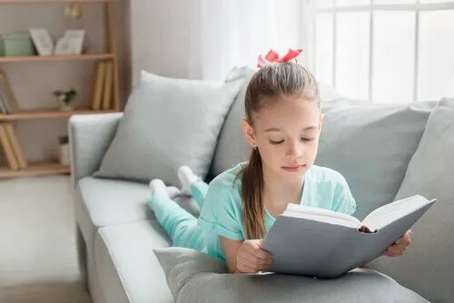 Niña tumbada en el sofá lee un libro.