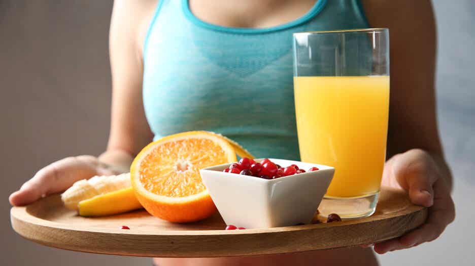 Mujer llevando una bandeja de comida con zumo, naranjas y un cuenco con frutos rojos tras hacer ejercicios.