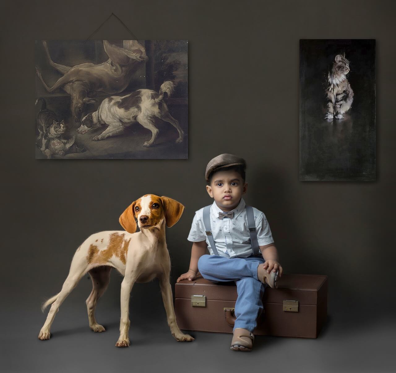 Niño sentado en una maleta acompañado de un perro con fondo oscuro y cuadros de animales.