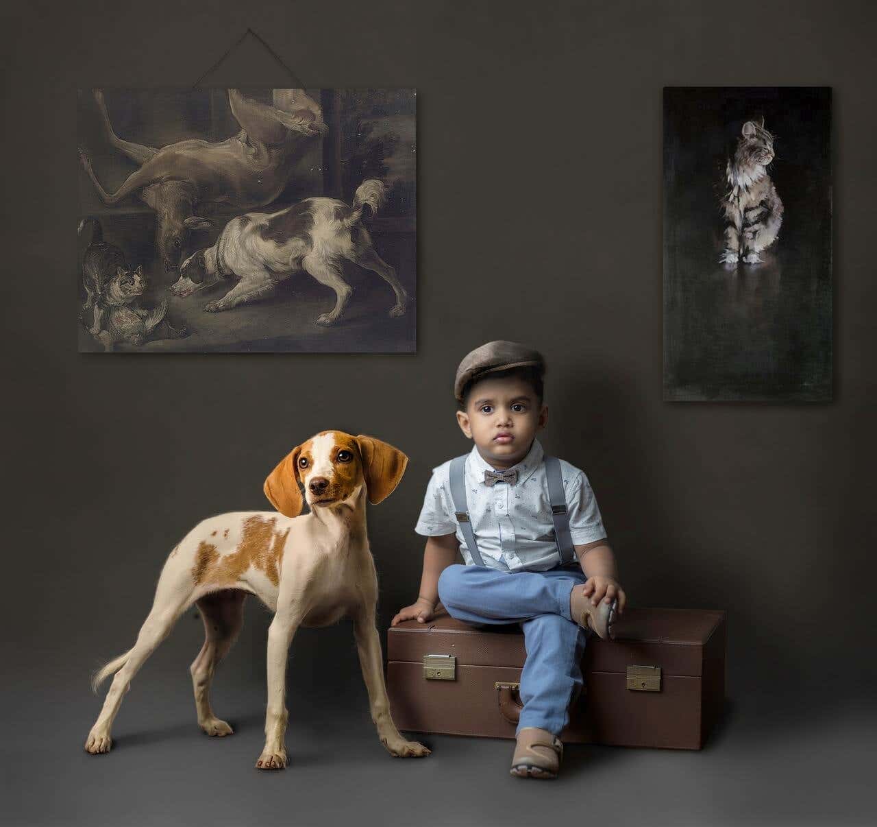 Niño sentado en una maleta acompañado de un perro con fondo oscuro y cuadros de animales.