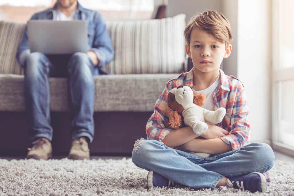 Niño sentado en la alfombra mientras su padre trabaja con el ordenador. Puede estar sufriendo maltrato.