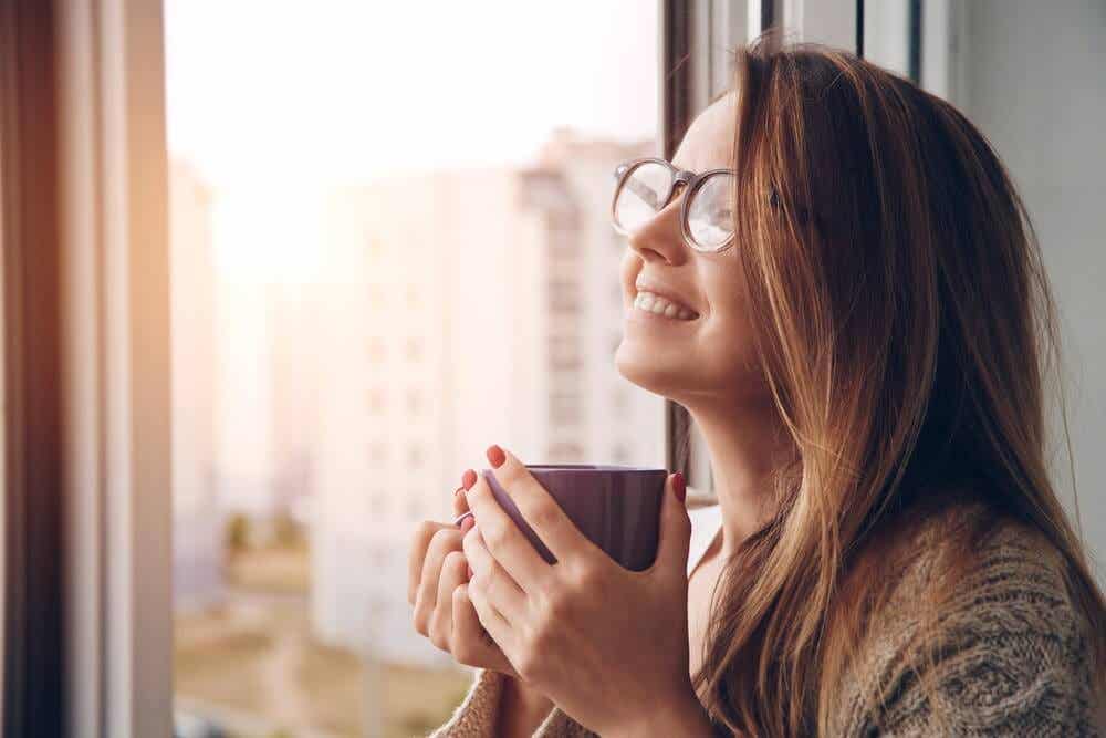 Mujer con una taza en la mano sonriendo y mirando por la ventana.