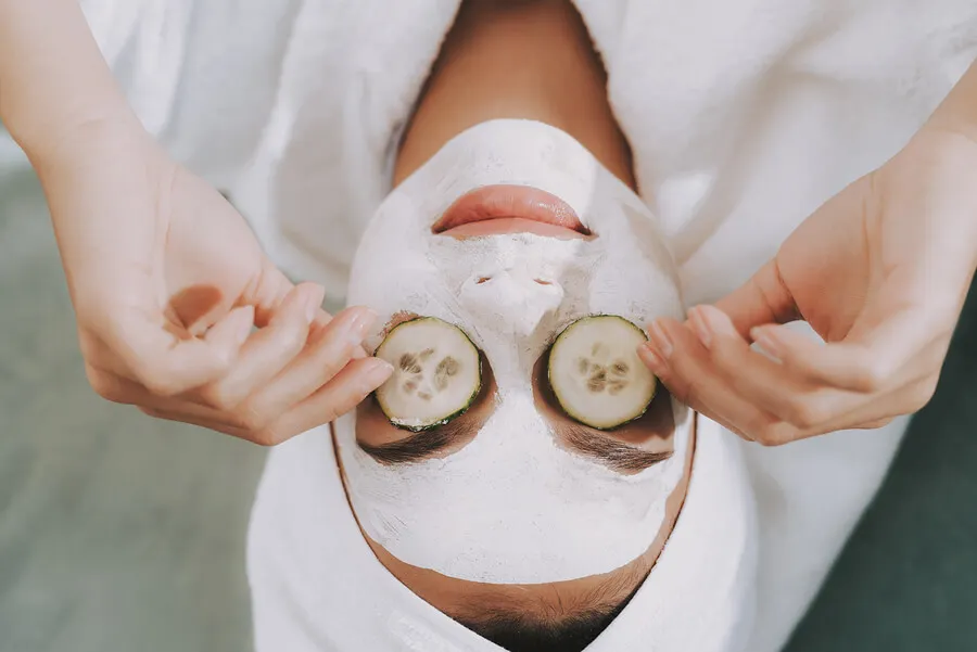 Mujer con mascarilla en la cara y pepino en los ojos.