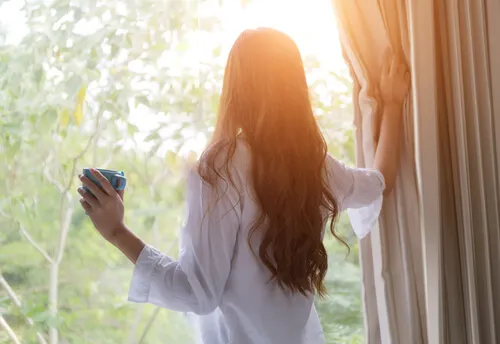 Mujer mirando por la ventana con café en mano.