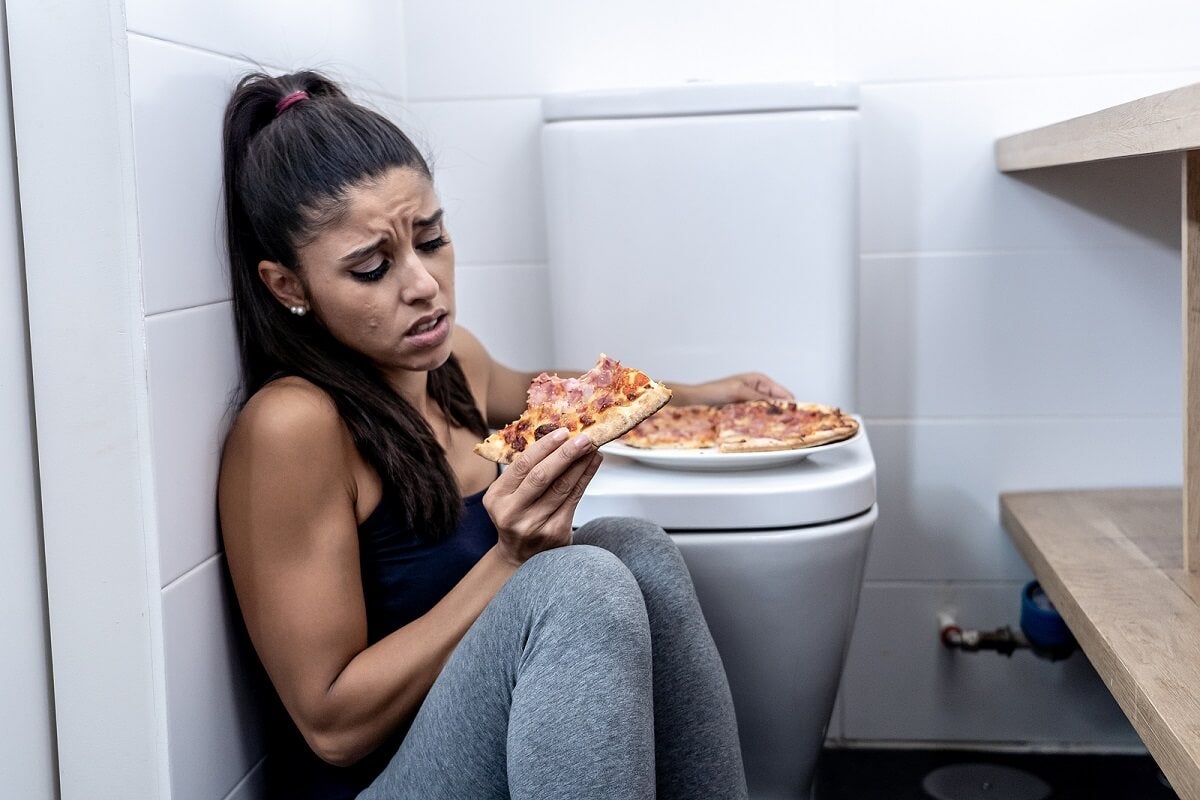 Schuldgefühle beim Essen - Frau isst eine Pizza im Badezimmer