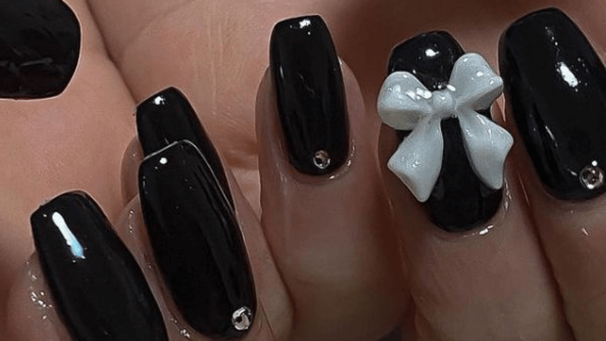 Uñas negras con brillantes y lazo blanco en el dedo anular.